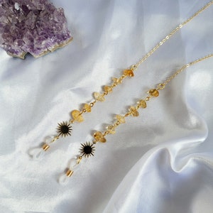 May include: Gold-colored eyeglass chains with yellow gemstone beads and black sunburst charms. The chains are displayed on a white satin surface, with a purple amethyst crystal in the background.