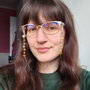 May include: A woman wearing gold-rimmed eyeglasses with a decorative chain. The chain features a sunburst charm, yellow gemstones, and a delicate gold chain. The glasses have a light blue tint.