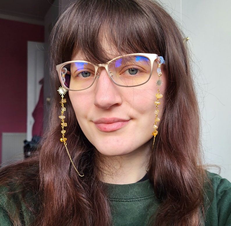 May include: A woman wearing gold-rimmed eyeglasses with a decorative chain. The chain features a sunburst charm, yellow gemstones, and a delicate gold chain. The glasses have a light blue tint.