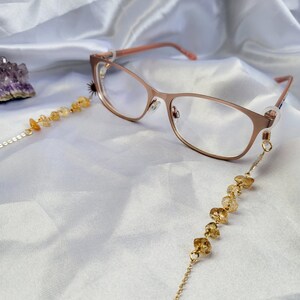 May include: Rose gold eyeglasses with a citrine beaded chain. The glasses have a delicate frame and clear lenses. The chain is made of small, golden-yellow citrine beads strung on a fine gold chain. The background is a white, silky fabric.