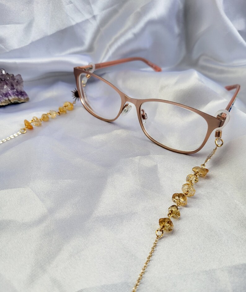 May include: Rose gold eyeglasses with a citrine beaded chain. The glasses have a delicate frame and clear lenses. The chain is made of small, golden-yellow citrine beads strung on a fine gold chain. The background is a white, silky fabric.