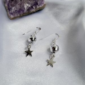 May include: A pair of silver earrings featuring a disco ball-shaped bead and a star charm. The earrings hang from a silver hook. The background is a white, silky fabric.