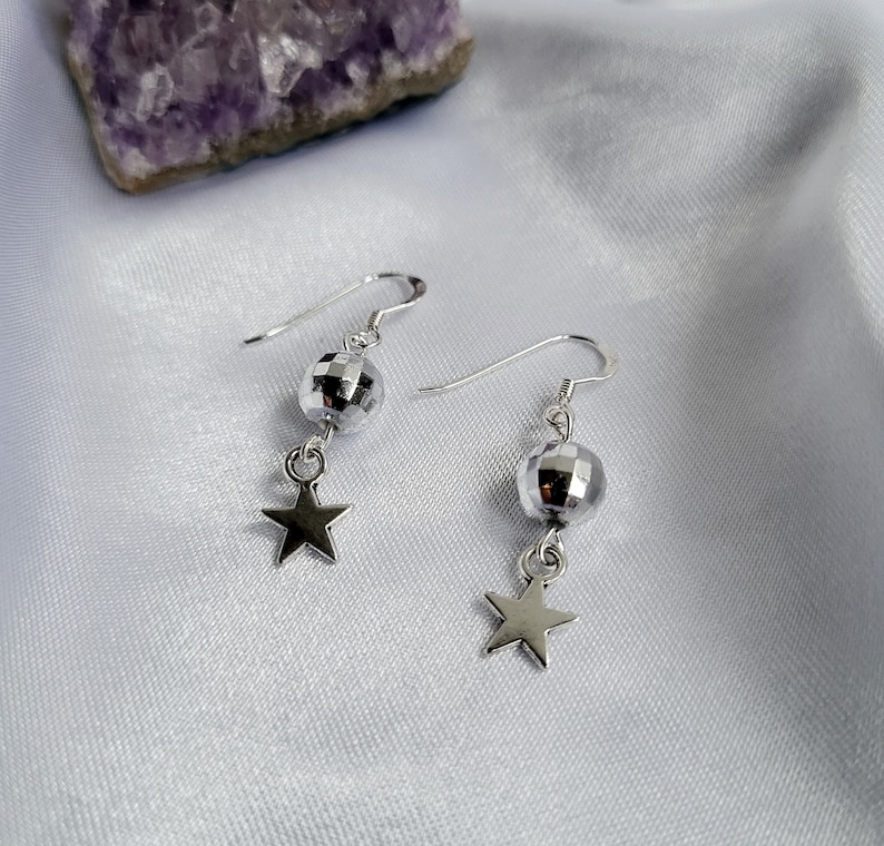May include: A pair of silver earrings featuring a disco ball-shaped bead and a star charm. The earrings hang from a silver hook. The background is a white, silky fabric.