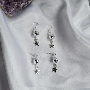 May include: Silver-toned earrings featuring a disco ball design with a dangling star charm. The earrings are available in both hook and stud styles. The jewelry is displayed on a white, silky fabric, with a purple crystal in the background.