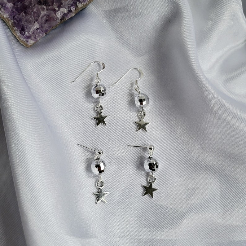 May include: Silver-toned earrings featuring a disco ball design with a dangling star charm. The earrings are available in both hook and stud styles. The jewelry is displayed on a white, silky fabric, with a purple crystal in the background.