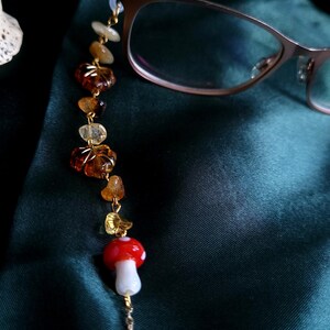 May include: Eyeglass chain with a whimsical design. The chain features a red and white mushroom charm, amber-colored beads, and a gold-colored chain. The glasses are a light brown color. The background is a dark green.