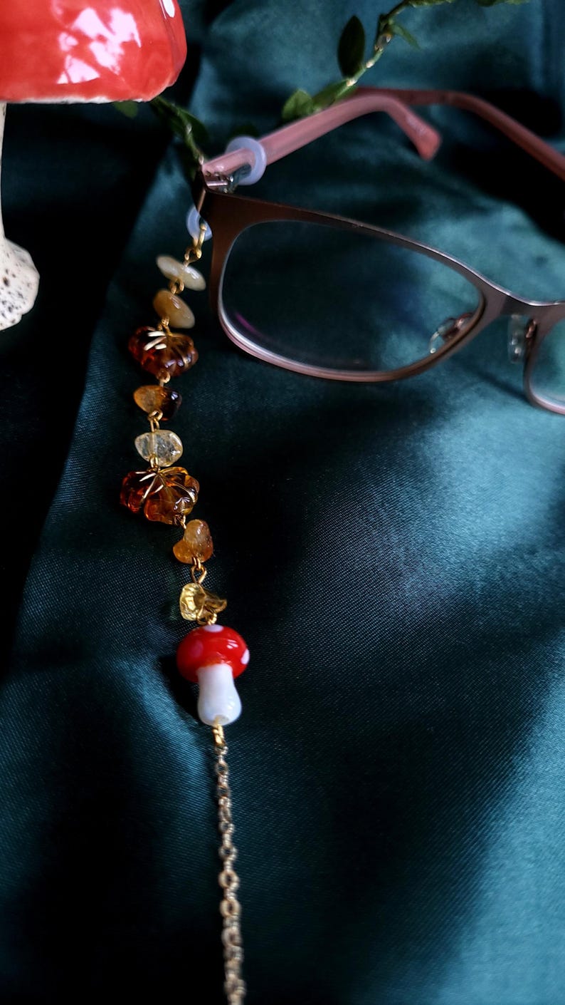May include: Eyeglass chain with a whimsical design. The chain features a red and white mushroom charm, amber-colored beads, and a gold-colored chain. The glasses are a light brown color. The background is a dark green.