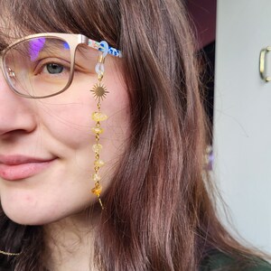 May include: A gold-colored eyeglass chain featuring a sunburst charm and a series of yellow-orange gemstone beads. The chain is attached to the glasses with clear loops. The glasses have a floral pattern on the arms.