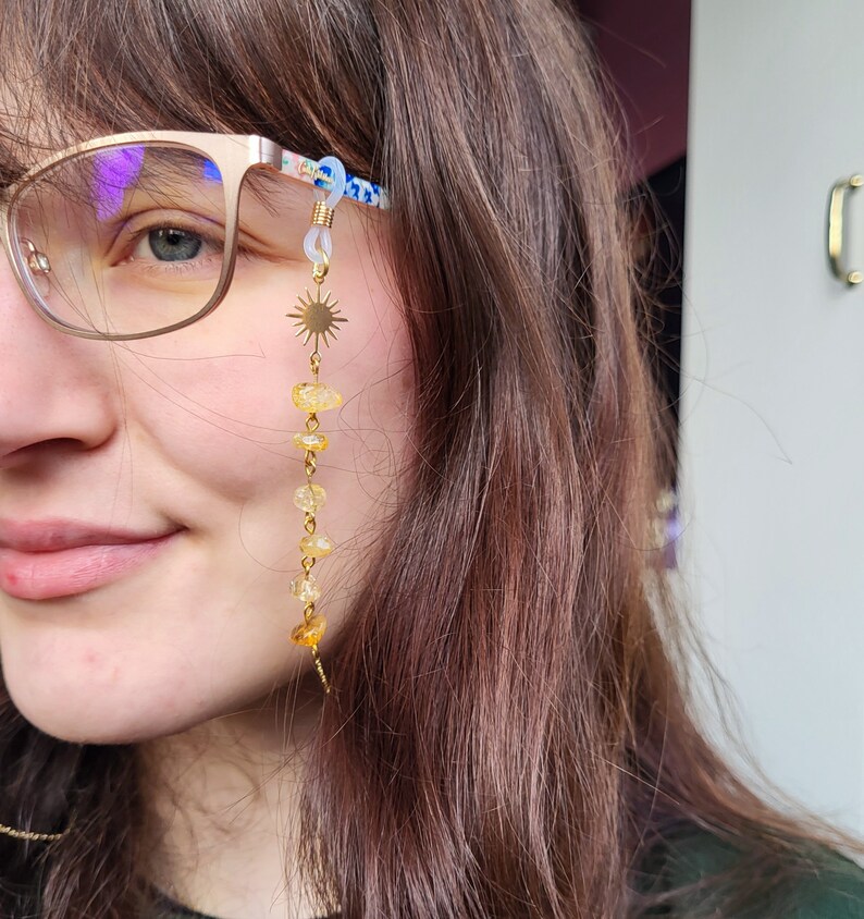 May include: A gold-colored eyeglass chain featuring a sunburst charm and a series of yellow-orange gemstone beads. The chain is attached to the glasses with clear loops. The glasses have a floral pattern on the arms.