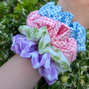 May include: A set of three fabric hair scrunchies featuring a gingham check pattern in light blue, pink, and a combination of light green and purple. The scrunchies are gathered with elastic, creating a ruffled, voluminous appearance on the fabric.