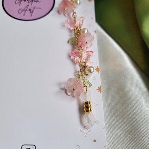 May include: An eyeglass chain with pink flower and butterfly charms, green leaves, and faux pearls. The gold-toned chain has a white loop for glasses. The card in the background has the text "Laura Gordon Art".