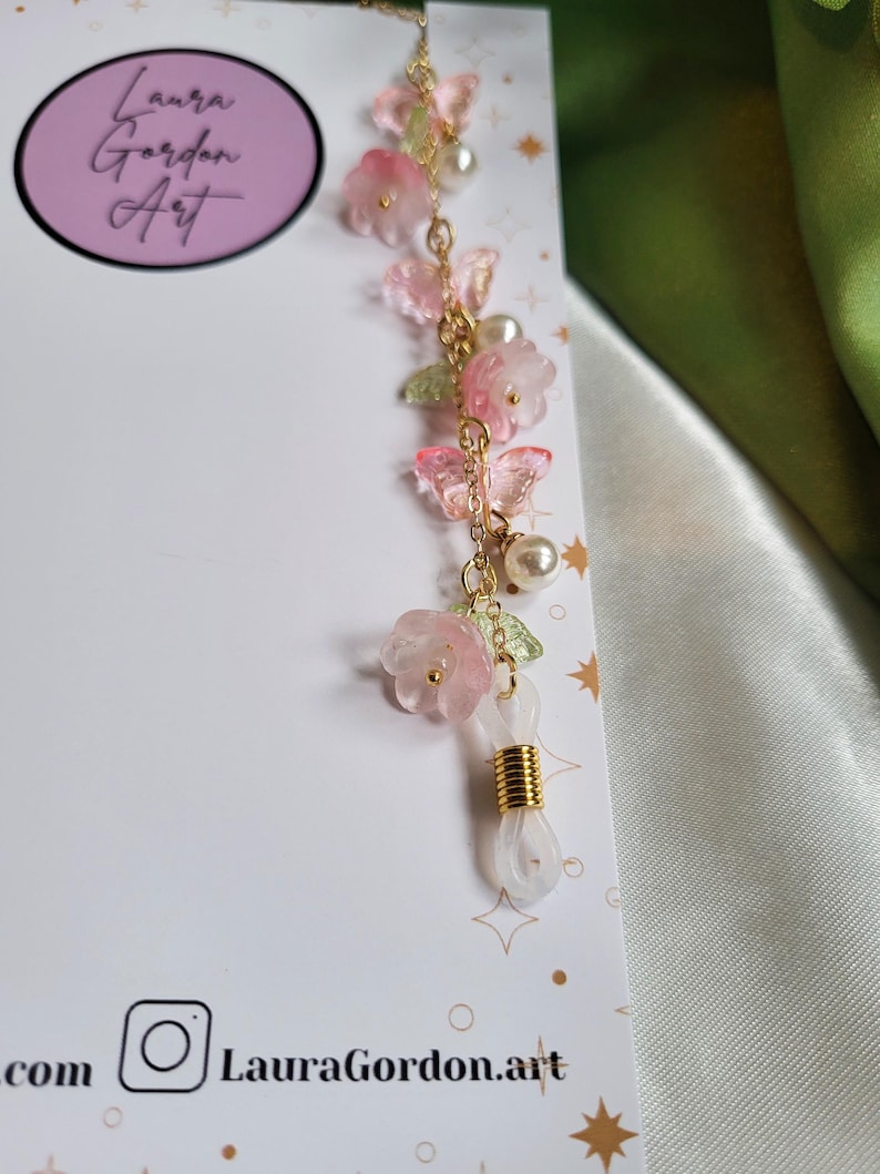 May include: An eyeglass chain with pink flower and butterfly charms, green leaves, and faux pearls. The gold-toned chain has a white loop for glasses. The card in the background has the text "Laura Gordon Art".