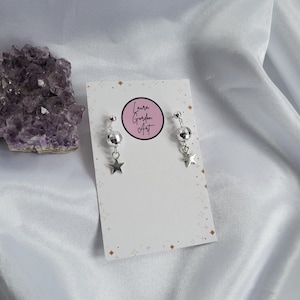 May include: Silver-toned earrings featuring a small disco ball and a star charm. The earrings are displayed on a white card with the text "Laura Gordon Art" in a pink circle. The background is a white, silky fabric.