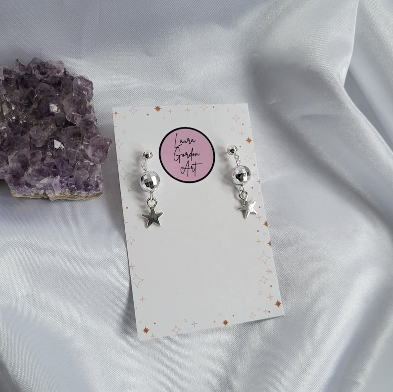 May include: Silver-toned earrings featuring a small disco ball and a star charm. The earrings are displayed on a white card with the text "Laura Gordon Art" in a pink circle. The background is a white, silky fabric.