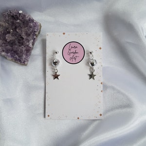 May include: Silver-toned earrings with a disco ball-shaped bead and a dangling star charm. The earrings are displayed on a white card with the text "Laura Gordon Art" in a pink circle. A purple crystal is in the background.