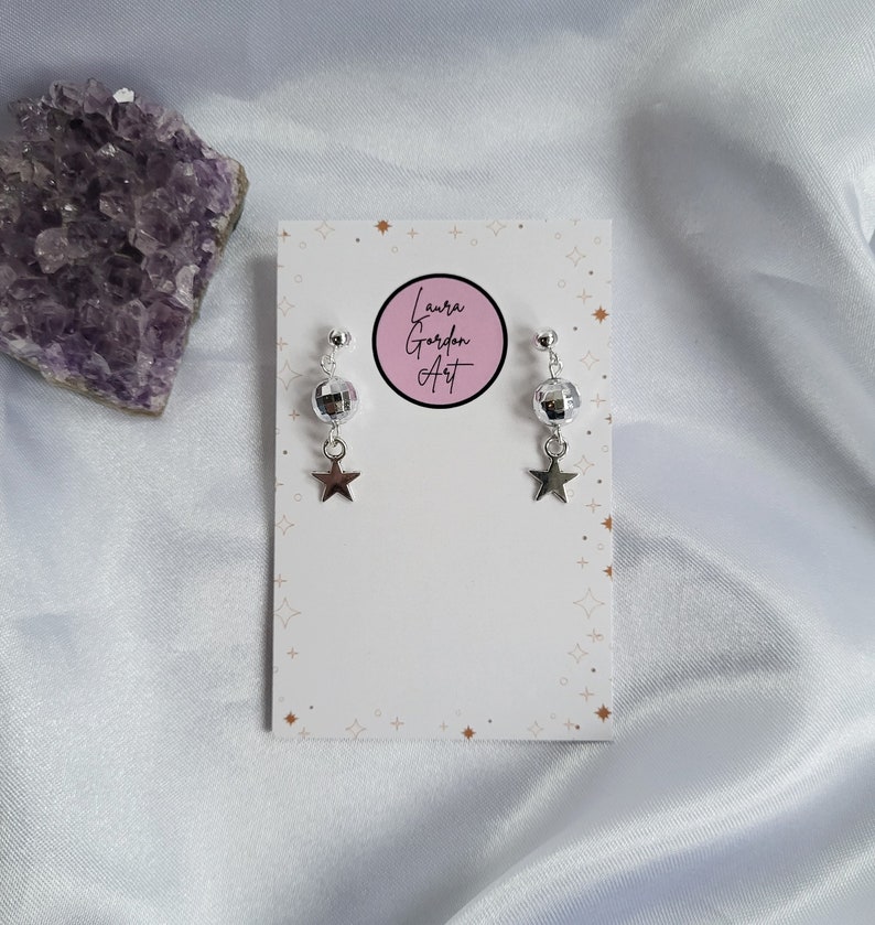 May include: Silver-toned earrings with a disco ball-shaped bead and a dangling star charm. The earrings are displayed on a white card with the text "Laura Gordon Art" in a pink circle. A purple crystal is in the background.