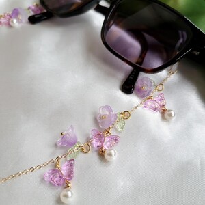 May include: Black sunglasses with a gold chain eyeglass holder. The chain features lavender flowers, translucent purple butterflies, small green leaves, and white pearls. The glasses are on a white satin surface.