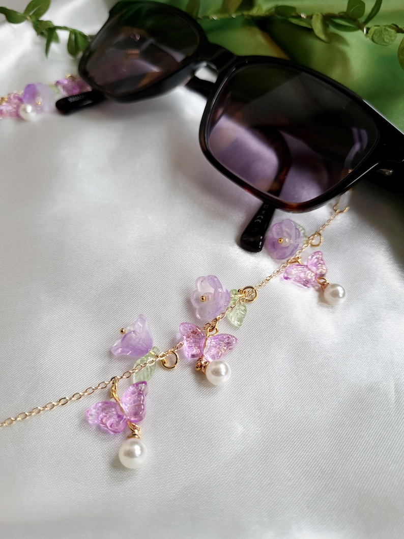 May include: Black sunglasses with a gold chain eyeglass holder. The chain features lavender flowers, translucent purple butterflies, small green leaves, and white pearls. The glasses are on a white satin surface.