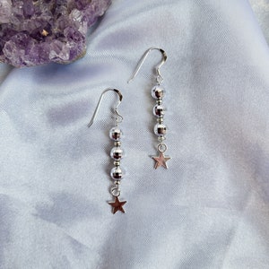 May include: A pair of silver dangle earrings. Each earring features a series of small, reflective beads and a small star charm at the bottom. The earrings are displayed on a light blue, silky fabric, with a purple amethyst crystal in the background.
