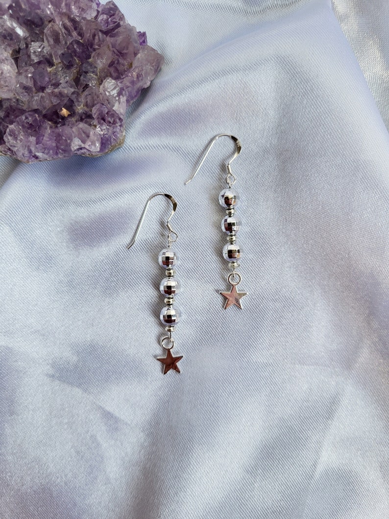May include: A pair of silver dangle earrings. Each earring features a series of small, reflective beads and a small star charm at the bottom. The earrings are displayed on a light blue, silky fabric, with a purple amethyst crystal in the background.
