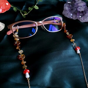 May include: Rose gold eyeglasses with a beaded chain featuring amber and clear stones, and red and white mushroom charms. The glasses rest on a dark green fabric, with a crystal and foliage in the background.