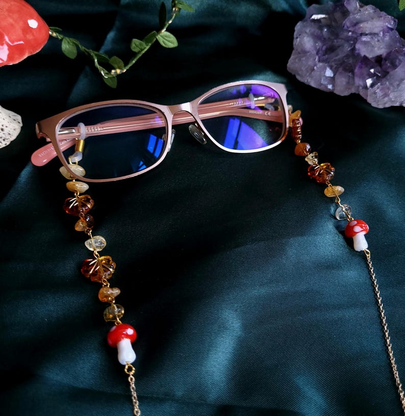 May include: Rose gold eyeglasses with a beaded chain featuring amber and clear stones, and red and white mushroom charms. The glasses rest on a dark green fabric, with a crystal and foliage in the background.