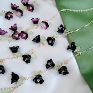 以下が含まれることがあります： 黒と紫の花のペンダントと緑の葉のアクセントが特徴の繊細なフローラルジュエリー。ペンダントは繊細なゴールドチェーンに通されています。背景は白と緑の生地のミックスです。