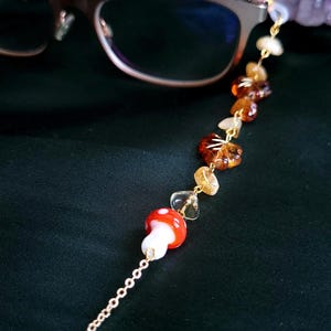 May include: Eyeglass chain with a gold-colored chain and decorative elements. The chain features a red and white mushroom charm, amber-colored beads, and clear crystal-like beads. The chain is designed to hold eyeglasses.