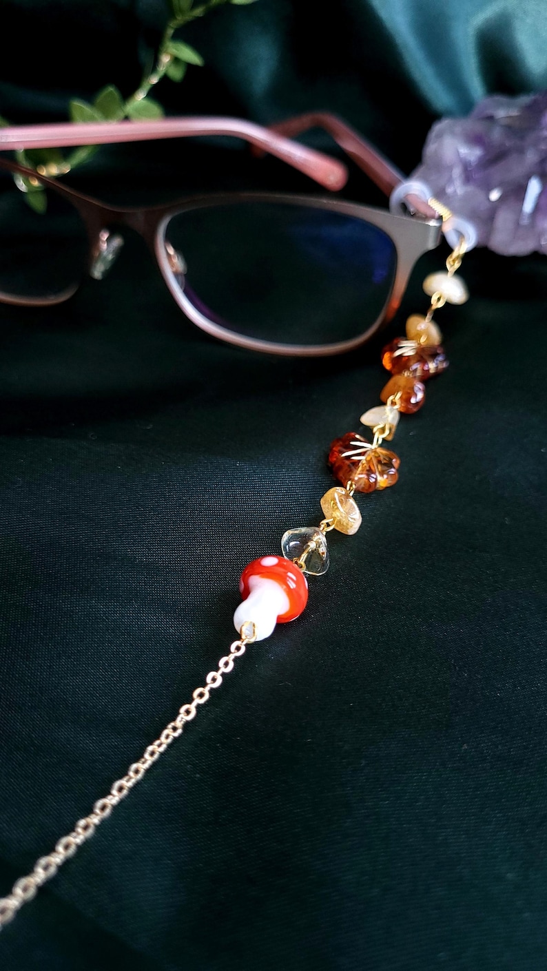 May include: Eyeglass chain with a gold-colored chain and decorative elements. The chain features a red and white mushroom charm, amber-colored beads, and clear crystal-like beads. The chain is designed to hold eyeglasses.