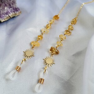 May include: Two gold-colored eyeglass chains with citrine gemstone beads and sunburst charms. The chains have white rubber loops for attaching to glasses. The background is a white, silky fabric with a purple crystal.