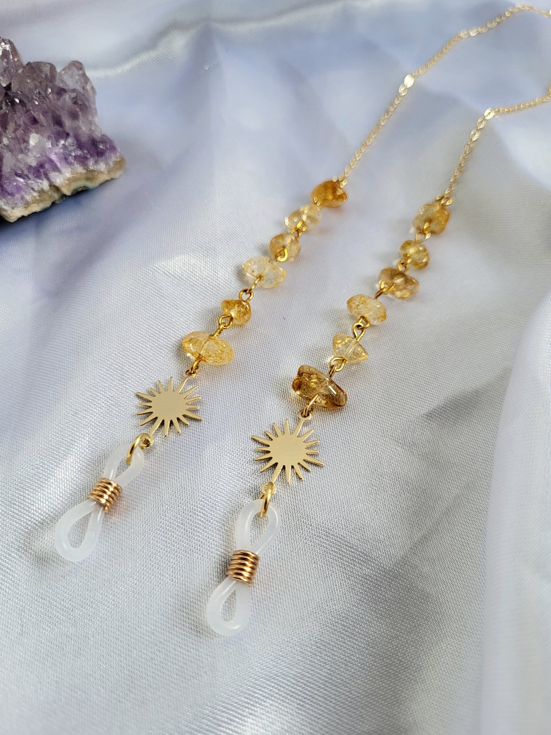 May include: Two gold-colored eyeglass chains with citrine gemstone beads and sunburst charms. The chains have white rubber loops for attaching to glasses. The background is a white, silky fabric with a purple crystal.