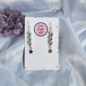 May include: Silver dangle earrings with a star charm. Each earring features a series of silver beads and a star-shaped charm. The earrings are displayed on a white card with the text "Laura Gordon Art" in a pink circle.