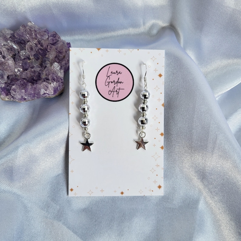 May include: Silver dangle earrings with a star charm. Each earring features a series of silver beads and a star-shaped charm. The earrings are displayed on a white card with the text "Laura Gordon Art" in a pink circle.