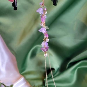 May include: A gold-coloured glasses chain with a delicate floral design. The chain features purple flower and butterfly charms, along with small pearls and green leaf accents. The chain is attached to black sunglasses.