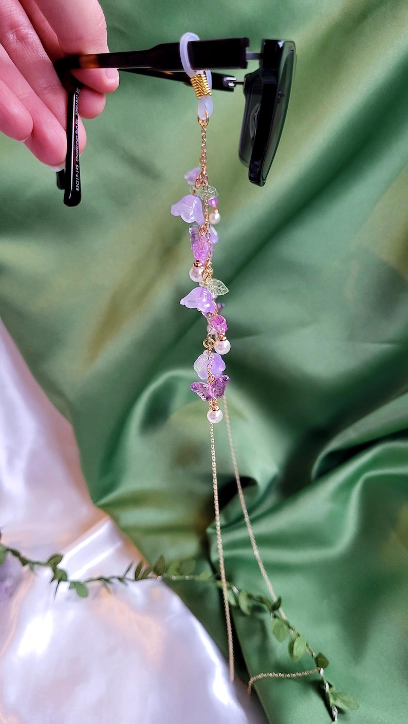 May include: A gold-coloured glasses chain with a delicate floral design. The chain features purple flower and butterfly charms, along with small pearls and green leaf accents. The chain is attached to black sunglasses.