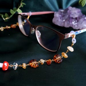 May include: Eyeglasses with a brown frame and a decorative eyeglass chain. The chain features a gold-colored chain with leaf-shaped amber beads, clear and yellow stones, and a red and white mushroom charm.