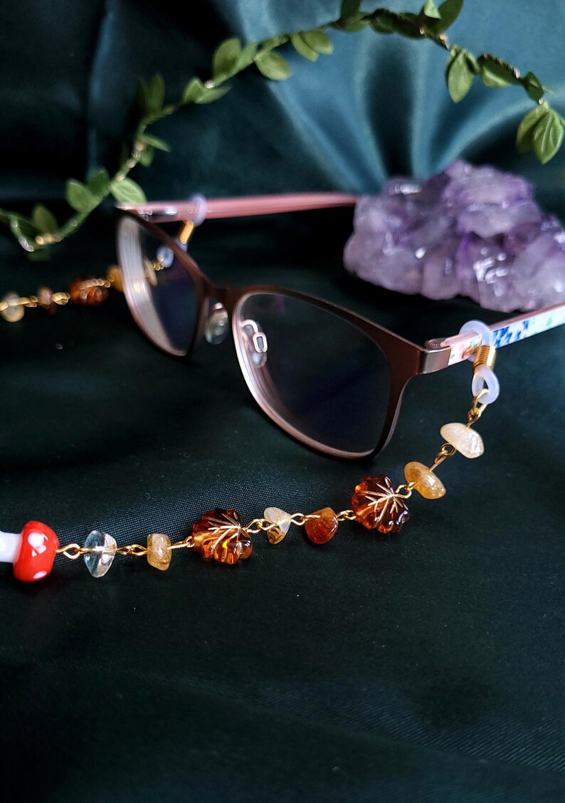 May include: Eyeglasses with a brown frame and a decorative eyeglass chain. The chain features a gold-colored chain with leaf-shaped amber beads, clear and yellow stones, and a red and white mushroom charm.