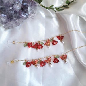 May include: Two gold-coloured eyeglass chains, each adorned with red flower and butterfly charms, green leaves, and faux pearls. They lie on a white satin surface. A purple crystal and greenery are in the background.