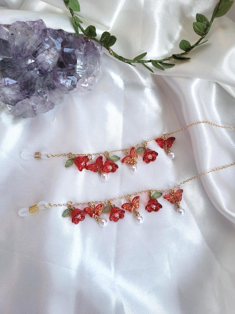 May include: Two gold-coloured eyeglass chains, each adorned with red flower and butterfly charms, green leaves, and faux pearls. They lie on a white satin surface. A purple crystal and greenery are in the background.