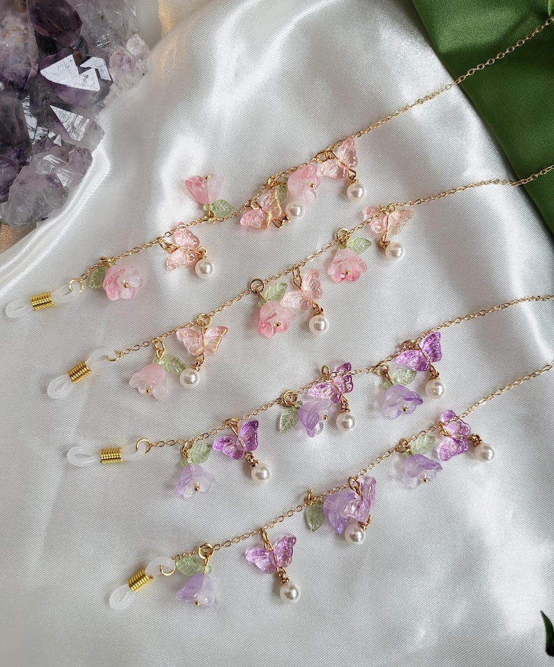 May include: Gold-coloured eyeglass chains adorned with floral and butterfly motifs. The chains feature pink, purple, and clear flowers, butterflies, and faux pearls. Clear rubber loops are attached for securing to glasses.