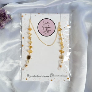 May include: A gold-colored eyeglass chain with yellow gemstone beads and sunburst charms. The chain is attached to a white card with the text "Laura Gordon Art" and social media handles. The chain is packaged in a clear plastic sleeve.