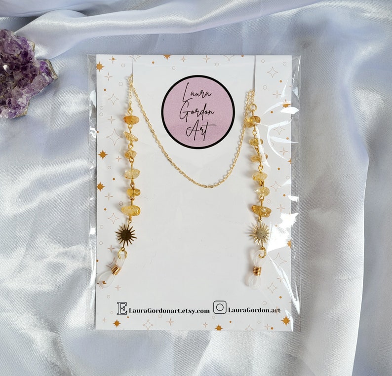 May include: A gold-colored eyeglass chain with yellow gemstone beads and sunburst charms. The chain is attached to a white card with the text "Laura Gordon Art" and social media handles. The chain is packaged in a clear plastic sleeve.