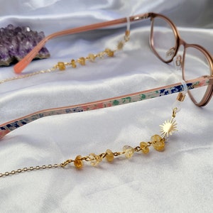 May include: Rose gold eyeglasses with a floral pattern on the temples and a gold chain eyeglass holder. The chain features yellow gemstone beads and a gold sunburst charm. The background is a white satin fabric.