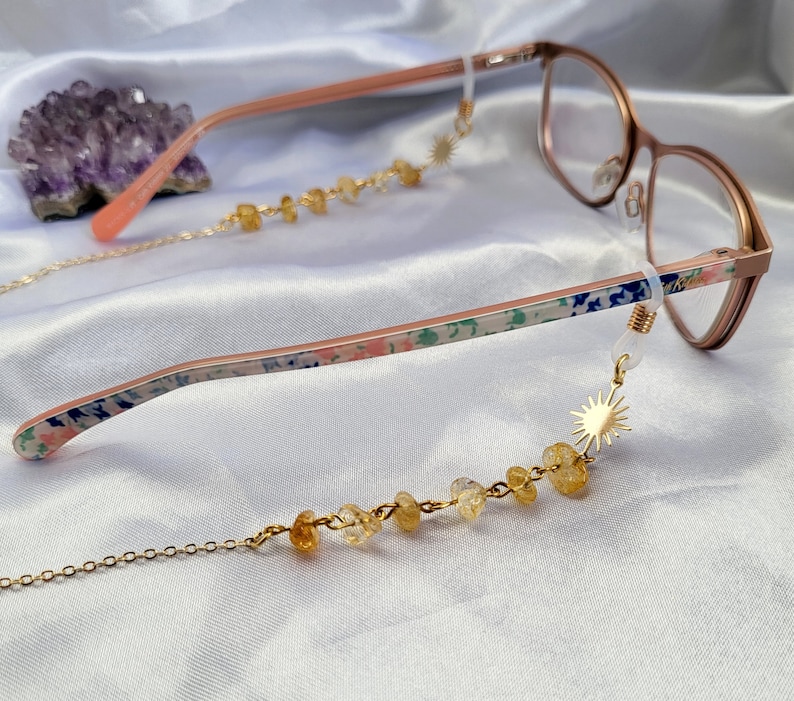 May include: Rose gold eyeglasses with a floral pattern on the temples and a gold chain eyeglass holder. The chain features yellow gemstone beads and a gold sunburst charm. The background is a white satin fabric.