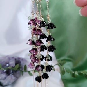 以下が含まれることがあります： ゴールドカラーのチェーンと黒と紫の花のビーズが付いたメガネチェーン。チェーンは透明なゴム製のループでメガネに取り付けられています。花は濃い紫と黒の混合で、小さな緑の葉のアクセントが付いています。