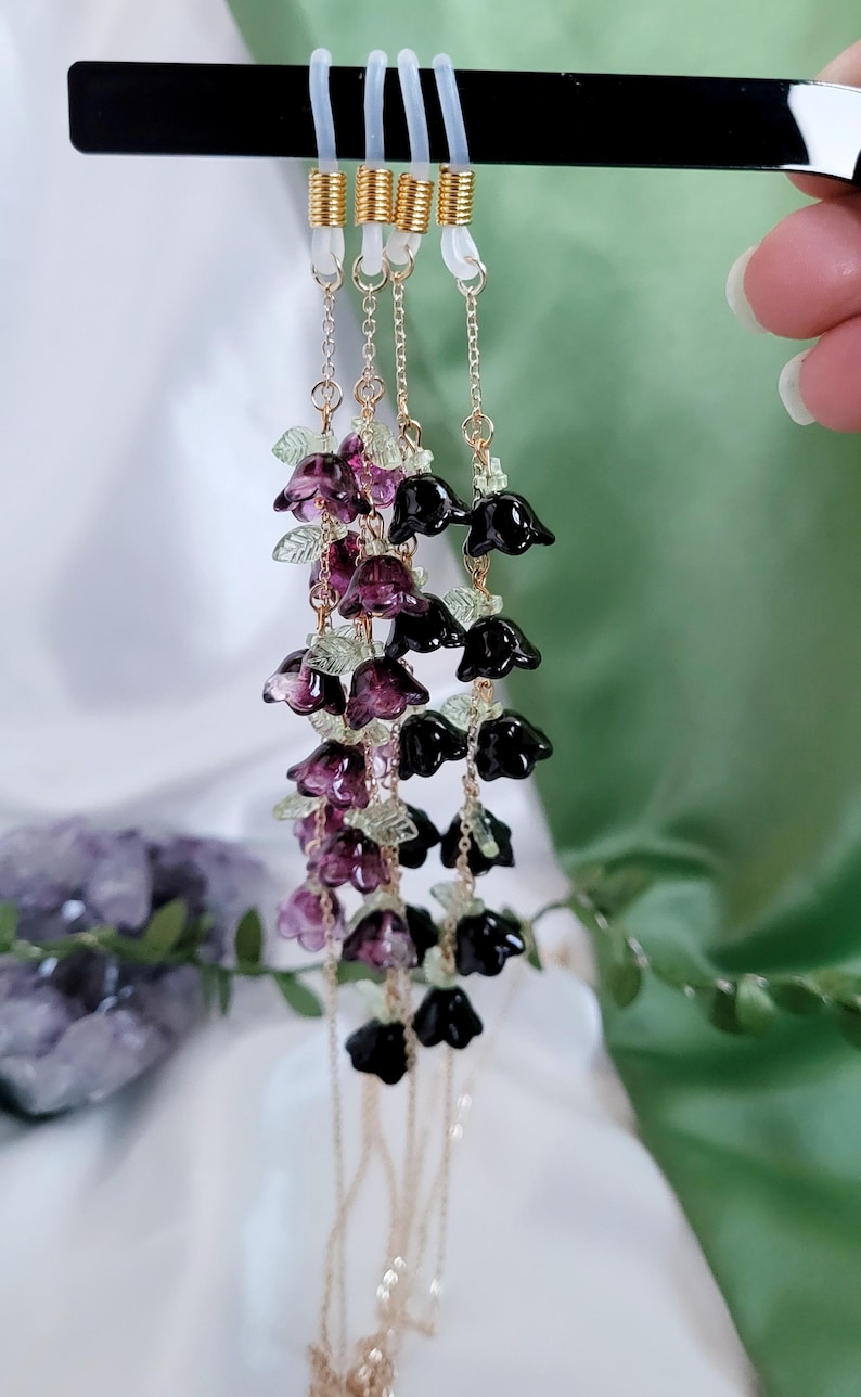 以下が含まれることがあります： ゴールドカラーのチェーンと黒と紫の花のビーズが付いたメガネチェーン。チェーンは透明なゴム製のループでメガネに取り付けられています。花は濃い紫と黒の混合で、小さな緑の葉のアクセントが付いています。