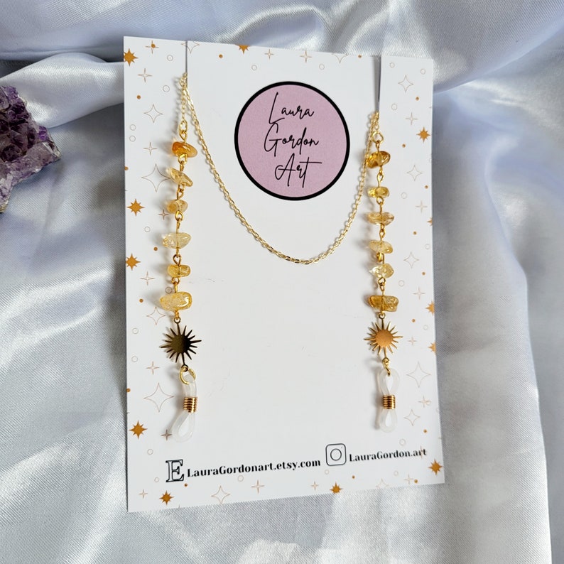 May include: Gold-colored eyeglass chain with citrine gemstone beads and sunburst charms. The chain is displayed on a white card with the text "Laura Gordon Art" and social media information.