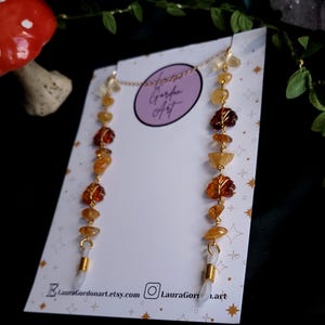 May include: A gold-colored eyeglass chain featuring a series of amber and yellow gemstone beads, including leaf-shaped accents. The chain is attached to white, plastic eyeglass holders. The background includes a white card with text and a dark, blurred backdrop.