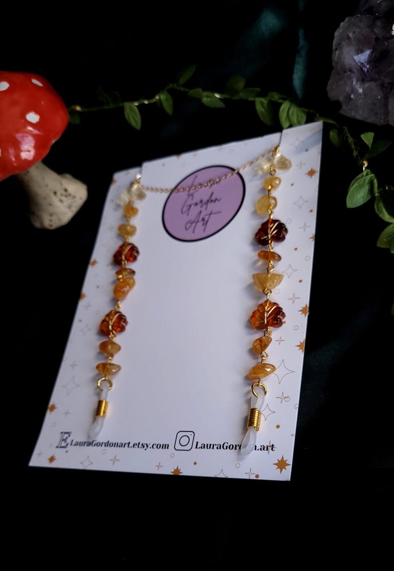 May include: A gold-colored eyeglass chain featuring a series of amber and yellow gemstone beads, including leaf-shaped accents. The chain is attached to white, plastic eyeglass holders. The background includes a white card with text and a dark, blurred backdrop.