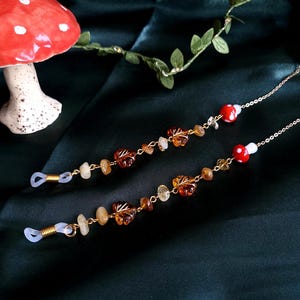 May include: Two eyeglass chains with gold-colored chains, amber-colored leaf charms, and small, round, red and white mushroom charms. The chains also feature small, light-colored stone beads. The background is a dark green fabric.